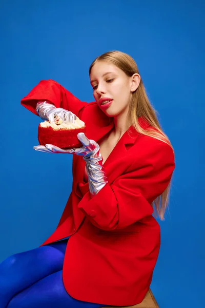 Model on diet. Young girl in unusual, freaky, bright outfit, wearing blue tights, red jacket holding birthday cake and going to eat with hands. Concept of high fashion, style and glamour, beauty, ad