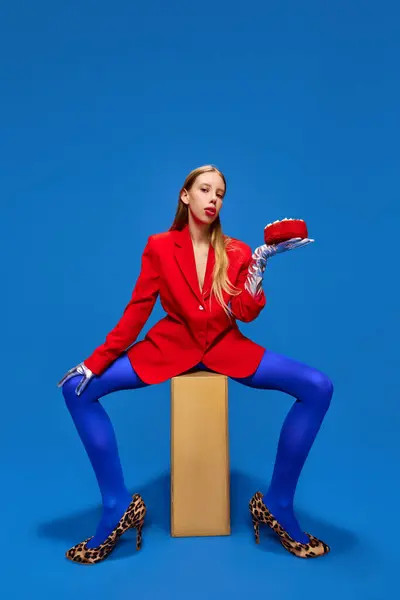 Fashion standards. Young lady posing in unusual, freaky, bright outfit, wearing blue tights, red jacket and animal printed heels holding birthday cake. Concept of high fashion, style, glamour, beauty.