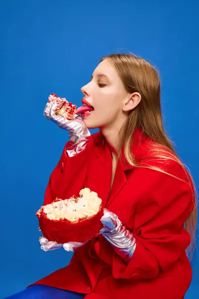 Portrait of overjoyed, freaky, unusual, vivid dressed girl sitting and licks broken off sweet and tasty big piece of cake against blue background. Concept of high fashion, style and glamour, beauty.