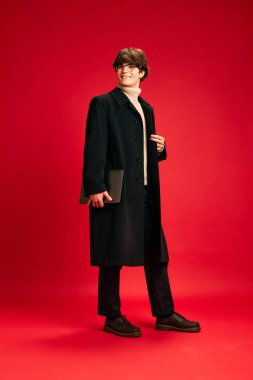 Genç çalışan. Başarılı bir gencin portresi. Sıradan giyinmiş ve dizüstü bilgisayarı kırmızı stüdyo arka planında tutuyor. İnsani duygular, güzellik, moda ve tarz kavramı.