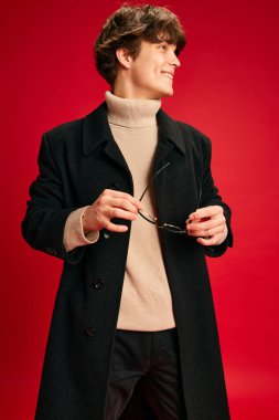 Gülümseyen genç adamın profil portresi modern ve sıcak kıyafetler giymiş kırmızı stüdyo arka planında gözlük tutuyordu. İnsani duygular, güzellik, moda ve tarz kavramı, samimi sonbahar.