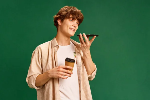Young attractive man, student talking on phone, making order and drinking refreshing hot coffee against green studio background. Concept of human emotions, facial expression, business, communication.