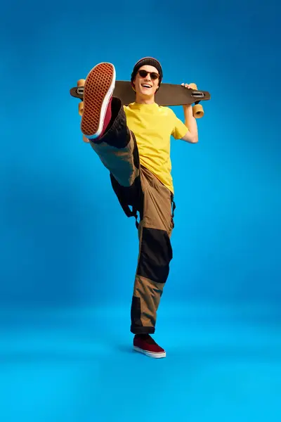 Full length portrait of cool dude rastaman, skateboarder holding skateboard and posing against blue studio background. Concept of youth, human emotions, self-expression, subcultures.