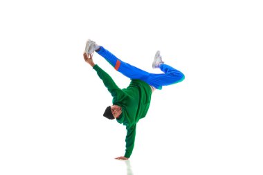 Genç, esnek sporcu adam, yeşil ve mavi üniformalı breakdance dansını beyaz stüdyo arka planına karşı yapıyor. Eylem kavramı, sanat, güzellik, spor, gençlik. Dansçı breakdance figürleri gösteriyor