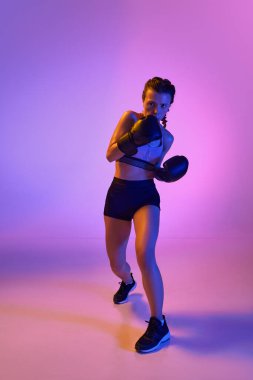 Dinamik uzunluktaki sporcu, kadın boksör, neon filtredeki değişken menekşe renk stüdyosuna karşı güç ve güven yayıyor. Spor anlayışı, yaşam tarzı, sağlık, rekabet.