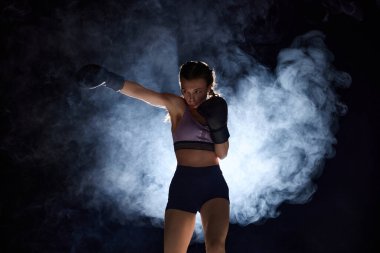 Sportif ve güçlü genç bir kadının dinamik portresi sahne dumanında siyah arka planda boks eğitimi alıyor. Spor anlayışı, aktif yaşam tarzı, sağlık, rekabet.