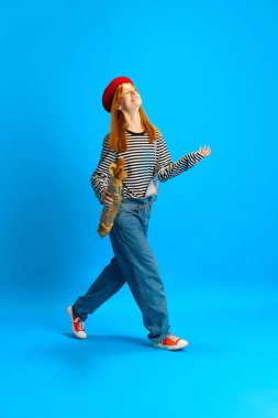 Mutlu neşeli kadın, kırmızı bereli retro moda kıyafeti giymiş mavi stüdyo arka planında baget ekmeğiyle yürüyor. İnsan duyguları kavramı, moda, tarz, satış sezonu, gençlik kültürü.
