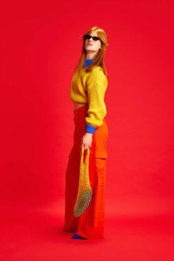 Eski moda parlak kıyafetler giymiş ve canlı kırmızı arka planda poz veren çekici bir genç kızın tam boy portresi. Modern moda kavramı, parlak yaşam tarzı, tarz eğilimleri, sonbahar, sonbahar