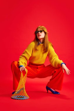 Havalı, çekici bir kız, eski moda kıyafetlerle poz veriyor ve kırmızı arka planda şeffaf bir çanta tutuyor. İnsan duyguları kavramı, modern moda, tarz eğilimleri.