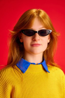 Kızıl saçlı, kendine güvenen genç bir kızın kırmızı arka planda güneş gözlüğüyle kameraya bakışının yakın portresi. İnsan duyguları kavramı, modern moda, parlak hayat, yaşam tarzı, tarz eğilimleri.