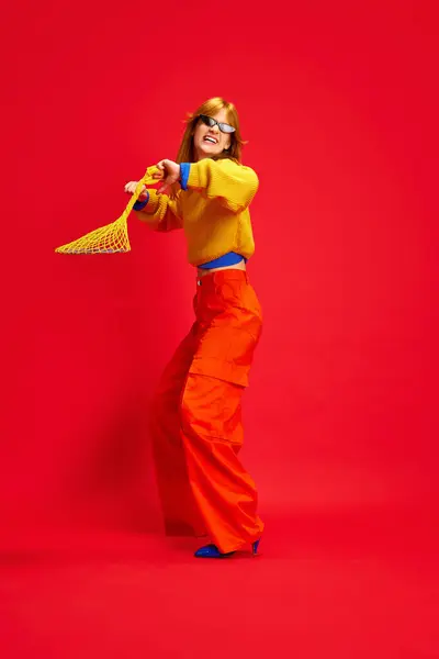 Creative portrait of funny girl, dancing dressed bright retro fashion outfit with sunglasses, accessories against vivid red background. Concept of human emotions, modern fashion, bright life, trends.