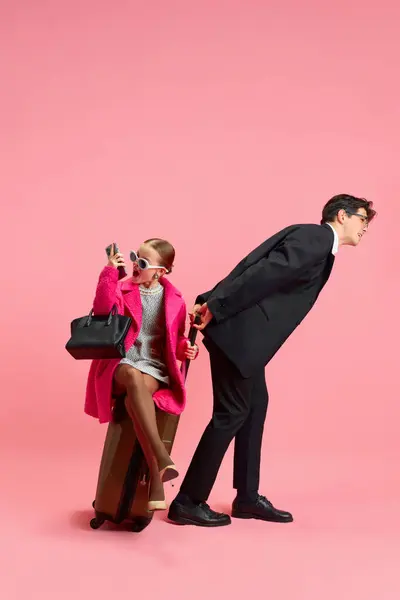 stock image Schoolgirl dressed in lady boss outfit sitting on bag and her personal assistant trying to pulling it against pink studio background. Concept of business, office, work, job, occupation, management.