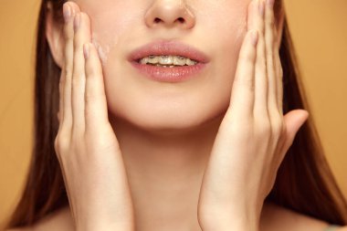 Bej arka planda sağlıklı ve ışıl ışıl görünmesini sağlamak için cildine krem süren genç bir kadının kesilmiş portresi. Doğal güzellik ve makyaj anlayışı, cilt bakımı prosedürleri.