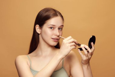 Uzun esmer saçlı çekici bir kadının portresi kameraya bakıyor ve bej arka planda kozmetik ürünler tutuyor. Doğal güzellik, makyaj, moda, tarz kavramı. İlan için alanı kopyala