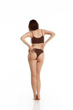 Arkadan bakınca, kahverengi iç çamaşırlı, ince ve esnek genç bir kadının beyaz arka planda zarif bir şekilde el kaldırdığı görülüyor. Kadın sağlığı, kadınlık, sağlık kavramı.