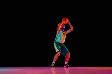 Hareket halindeki basketbol oyuncusunun tam boy portresi, karışık neon ışıkta siyah arkaplana karşı sanatın mükemmelleştirilmesine olan bağlılığı vurguluyor. Spor anlayışı, hobi, aktif yaşam tarzı. Ad