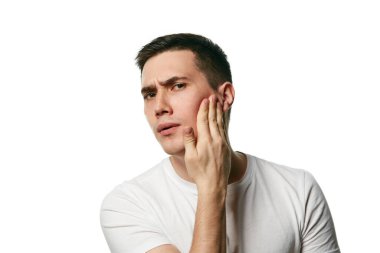 Genç yakışıklı adam şaşırtıcı bir şekilde kameraya bakıyor ve beyaz arka planda tenine dokunuyor. Güzellik ve hijyen kavramı, vücut bakımı, yaşlanma karşıtı prosedürler. Uzayı kopyala Ad