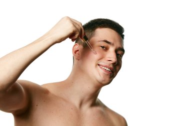 Kozmetik ürünler uygularken gülümseyen üstsüz bir gencin portresi, beyaz arka plana serumu nemlendiriyor. Güzellik tedavisi ve hijyen kavramı, vücut bakımı, yaşlanma karşıtı prosedürler.