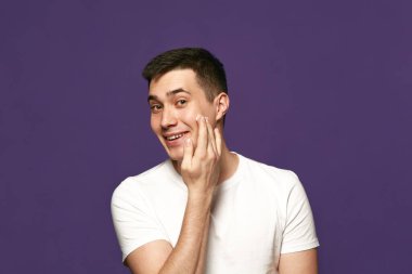 Cilt bakımı rutini. Yakışıklı genç adam kozmetik ürünler uyguluyor, mor arka plana nemlendirici krem sürüyor. Güzellik tedavisi ve hijyen kavramı, vücut bakımı, yaşlanma karşıtı prosedürler.