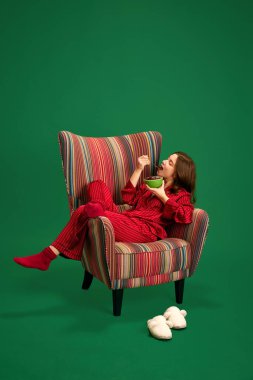 Kırmızı ipek pijamalarıyla retro koltuğa uzanmış güzel bir genç kızın portresi ve yeşil stüdyo arka planında lezzetli mısır gevreği yiyor. Mutlu yıllar kavramı, Mutlu Noeller.