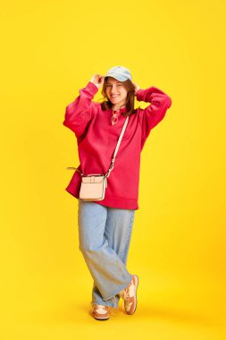 Mutlu, neşeli bir öğrencinin tam boy portresi, parlak sarı arka planda aşırı büyük kıyafetler giymiş bir turist. Moda ve tarz kavramı, seyahat, tatil, kültür, eğlence ve neşe.