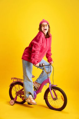 Neşeli, genç bayan, öğrenci gezgin kameraya bakıp gülümseyen ve kameraya bakan sarı stüdyo arka planında bisiklet süren. Moda ve tarz kavramı, seyahat, kültür.