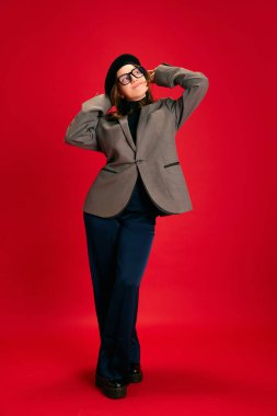Şık gözlüklü, şık giyinmiş, beresi kırmızı stüdyo arka planında olan zarif bir bayanın tam boy portresi. İş iletişimi, öğrenim ve bilgi kavramı.