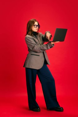 Genç zarif bir kadının tam boy portresi, kırmızı stüdyo arka planında dizüstü bilgisayarda çalışan gözlüklü bir öğrenci. İş iletişimi kavramı, online öğrenim ve bilgi, öğrenim.