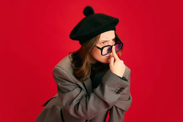 Portrait of young elegance lady adjusts his glasses and looks somewhere perplexed against red studio background. Concept of business, communication, study and knowledge, fashion and style.