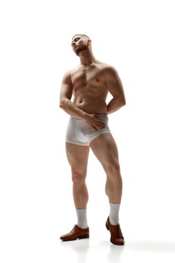 Beyaz stüdyo arka planında iç çamaşırlarıyla poz veren çekici ve kaslı çıplak bir adamın tam boy portresi. Erkek sağlığı, güzellik, vücut ve cilt bakımı, spor anlayışı. Vücut sanatı
