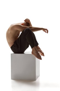 Çıplak gövdeli genç adam başını kucağına koydu ve kollarını beyaz arka planda küp üzerine oturttu. Erkek sağlığı, güzellik, vücut ve cilt bakımı, spor anlayışı. Vücut sanatı