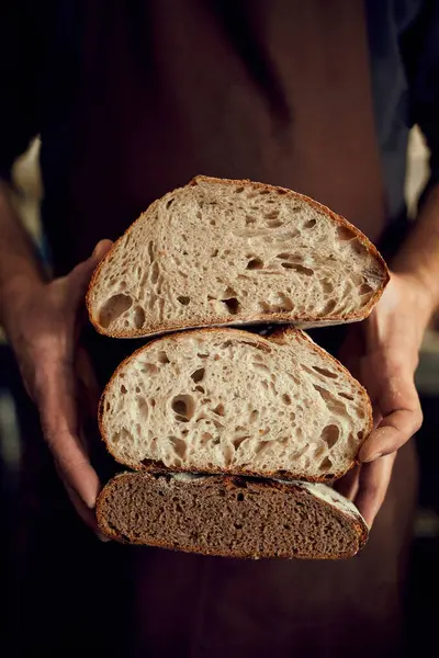 Bakery man, chef in brown apron holds in hands two halves of just whole grain hot loaf of bread with golden brown crispy crust. Concept of food, bakery, tradition and innovation, recipe.