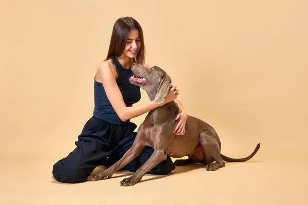 Look of love and tenderness. Young brunette woman sitting on floor and hugs with her favorite pet against beige studio background. Concept of animal, pet lover, friendship, companionship.