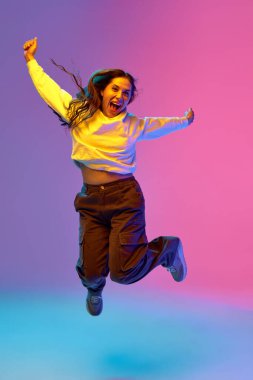 Full-length portrait of young positive woman jumping of joy against gradient purple-blue-pink background. Concept of beauty, youth, human emotions, self-expression, fun and happiness, fashion.