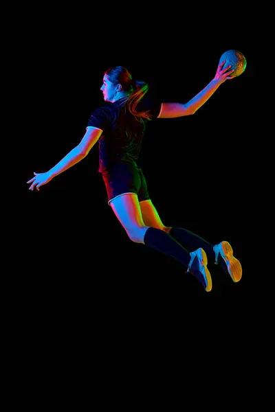 Determined woman, handball player in action, exhibiting focus and energy during training against black background in neon light. Concept of professional sport, hobby, movement, dynamic, championship.