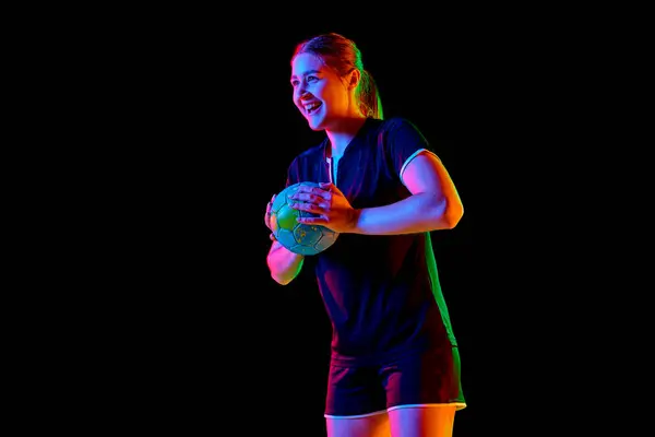 Portrait of young woman, handball player holding ball to make perfect precise serve early in game against black background in neon light. Concept of professional sport, championship 2024, victory. Ad