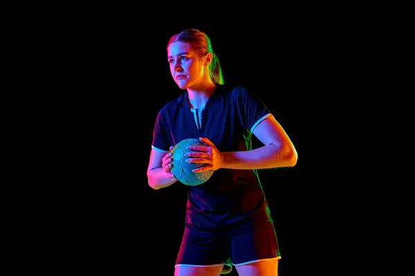 Portrait of focused woman, handball player holding ball to make perfect precise serve early in game against black background in neon light, filter. Concept of professional sport, championship 2024. Ad