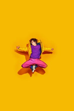 Canlı bir moda atlaması. Genç adam, şık, kalın kıyafetler giymiş, atlarken poz veriyor, parlak sarı arka planda özgürce hareket ediyor. Güzellik kavramı, modern tarz, alışveriş, satış sezonu.