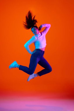 Neon ışıklarındaki pembe-turuncu arkaplana karşı havada poz veren aşırı mutlu genç kadının tam boy portresi. Kendini ifade etme, güzellik ve moda, enerji ve dinamik kavramı. Ad