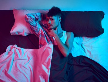 Beyond Etiketleri. Cinsiyetin akıcılığını kucaklamak. Genç erkek modelin çarpıcı fotoğrafı, düşünceli bir şekilde neon ışıkta kameraya bakıyor. Kendini ifade etme kavramı, erkeklik ve kadınlık, kimlik, gerçeküstü