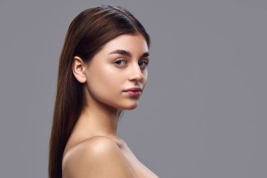 Açık tenli, doğal makyajlı ve omuzlarının üzerinden gri arka planda kameraya bakan genç bir kadının portresi. Doğal güzellik kavramı, cilt bakım ürünleri, besin takviyeleri.