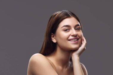 Çıplak omzuyla genç esmer kadın, elini uzatıp gri stüdyo arka planına bakan kameraya gülümsüyor. Besin takviyesi kavramı Vitamin ve mineral alımıyla temiz cilt.