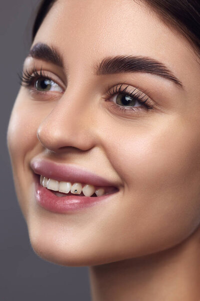 Крупный план портрета женского модельного лица с идеальными губами и макияжем глаз, улыбающимся на фоне серого студийного фона. Концепция продукта для укладки бровей, природная красота, полные брови.