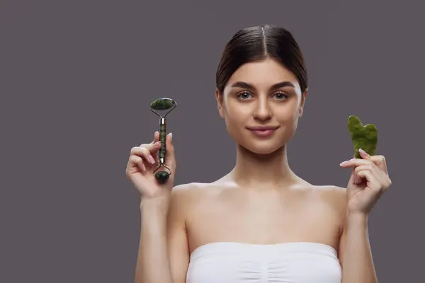 Naturally beautiful woman presenting roller and scraper from natural stone for facial massage against grey background. Concept of wellness, spa, facial treatment and anti-aging procedures.