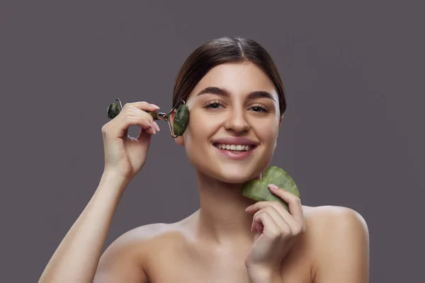 Naturally beautiful, positive young lady massaging her face with roller and scraper while looking at camera with smile against grey background. Concept of wellness, facial treatment and anti-aging.