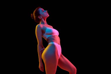 Beyaz iç çamaşırlı genç bir kadının, neon ışıkta siyah stüdyo arkaplanına bakarken yan görüntüsü. Sağlık, kilo kaybı, tedavi, sağlık, sağlık, sağlık, kaplıca kavramı.