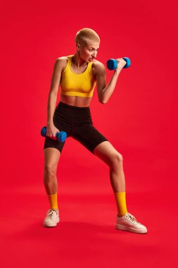 Dambılı eller için egzersiz yapan odaklanmış bir kadının dinamik portresi, sarı bluzlu, siyah şortlu, kırmızı stüdyo arka planına karşı. Spor, spor, aktif ve sağlıklı yaşam tarzı kavramı.