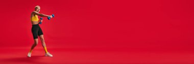 Pankart. Dambıllarla egzersiz yapan bir kadın. Kırmızı stüdyo arkaplanına karşı. Metninizi eklemek için negatif alanı olan. Spor anlayışı, fitness, aktif ve sağlıklı yaşam tarzı, motivasyon.