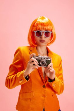 Vibrant fashion. Young woman in trendy sunglasses dressed orange blazer, with wig looking at camera and holds photo camera. Concept of personal expression and confidence through style. clipart