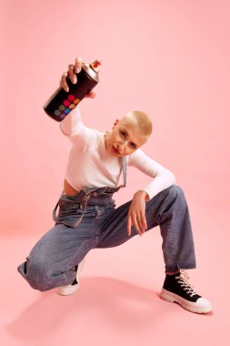 Genç kadın, kotlu modern sanatçı elinde boya şişesi tutuyor ve pembe arka planda kameraya duygusal olarak bakıyor. Gençlik, moda, şehir, kültür ve tarz kavramı.